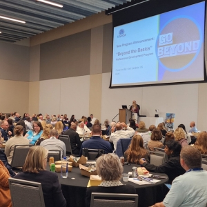 wide shot of a ballroom of conference attendees seated at tables listening to a presenter on stage with a screen overhead
